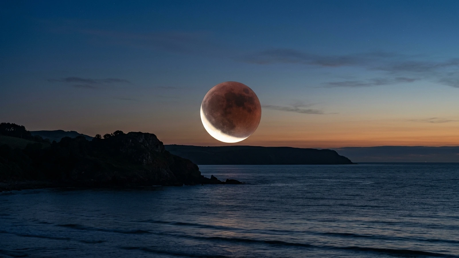 A deeply eclipsed copper-red Moon hanging low over a UK horizon in dawn twilight, with a thin bright sliver remaining on one edge