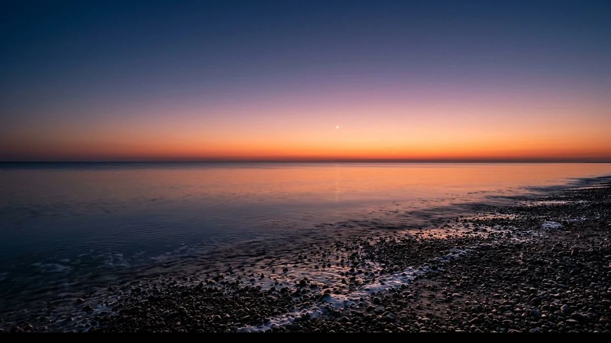 Brilliant Venus low in orange twilight over a flat sea horizon, with a very thin crescent Moon below it and to the right