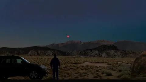 A digital rendering of a person standing in a desert landscape at night, with several red light sources visible in the sky.